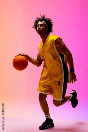 Image of biracial basketball player bouncing basketball on pink to orange background