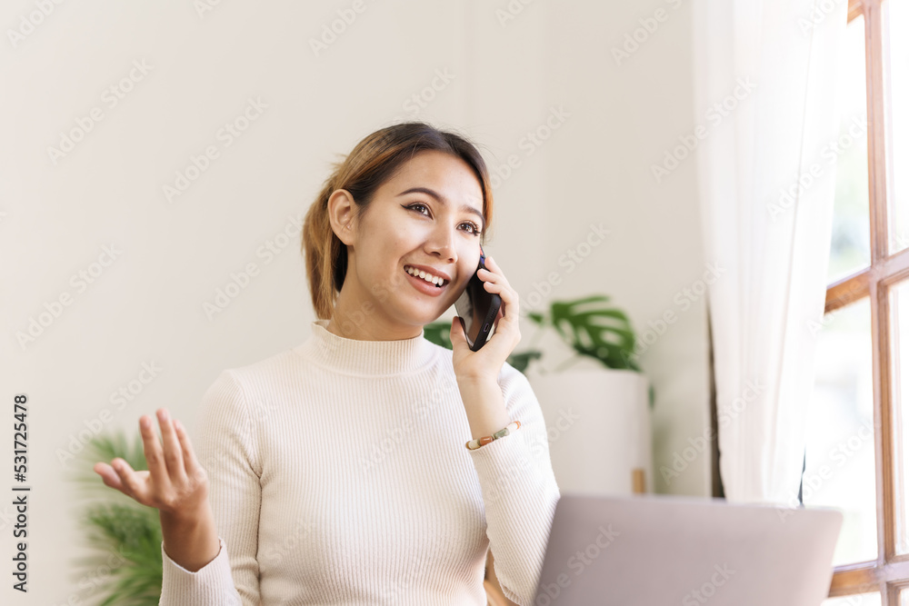 Happy asian businesswoman sitting at desk talking with mobile phone with somebody in office workplace.