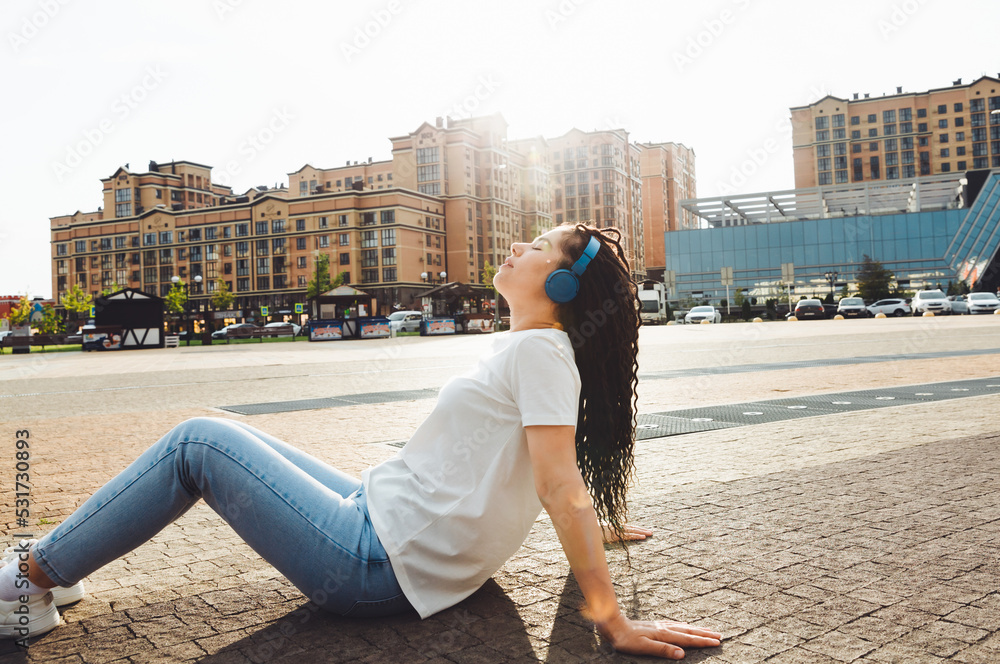 A smiling girl with dreadlocks in headphones is sitting on the floor in the park. A happy young woman relaxing with headphones, enjoying music. Space for copying.generation z