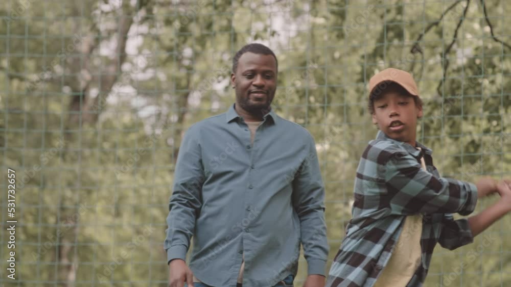 Medium slowmo of enthusiastic Black man teaching his teenage son to bat in baseball, practicing outdoors in park in summer Medium slowmo of enthusiastic Black man teaching his teenage son to bat in baseball, practicing outdoors in park in summer