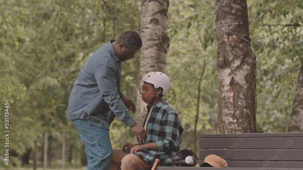 Slowmo of African American man helping his 12 year old son to put on safety equipment while having outdoor baseball training in park Slowmo of African American man helping his 12 year old son to put on safety equipment while having outdoor baseball training in park