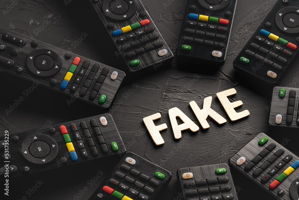 the word fake is assembled from wooden letters on a dark background, surrounded by TV remotes