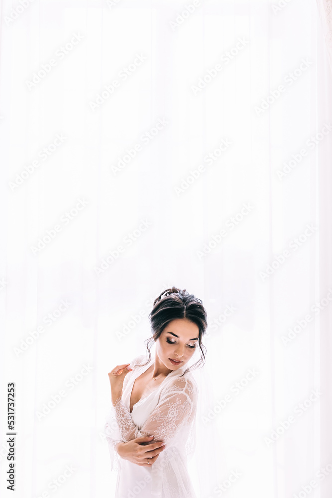 Morning of bride before the wedding. Beautiful young woman with long veil in a white robe and a negligee at the window. Morning of bride before the wedding. Beautiful young woman with long veil in a white robe and a negligee at the window.