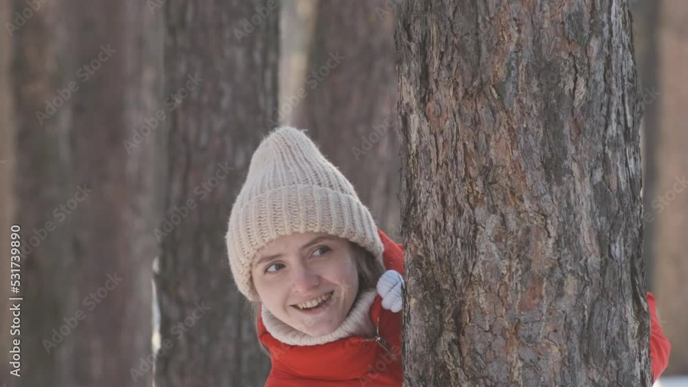 Young woman playfully peeks out from behind tree. Girl plays hide-and-seek in the woods in winter. Young woman playfully peeks out from behind tree. Girl plays hide-and-seek in the woods in winter.