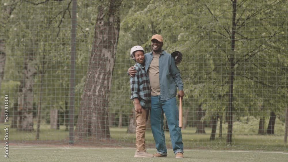 Full length slowmo portrait of cheerful African American father and his teenage son embracing each other and smiling at camera while standing on sports field in park during baseball training Full length slowmo portrait of cheerful African American father and his teenage son embracing each other and smiling at camera while standing on sports field in park during baseball training