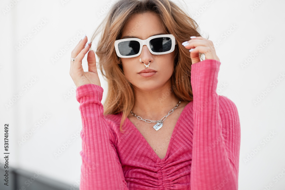 Fresh summer beautiful portrait of a pretty fashionable redhead woman with piercings with stylish cool white sunglasses in a pink knit top sweatshirt stand and posing near a white wall on the beach Fresh summer beautiful portrait of a pretty fashionable redhead woman with piercings with stylish cool white sunglasses in a pink knit top sweatshirt stand and posing near a white wall on the beach