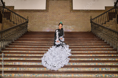 Young teenage woman in white dance suit with black polka dots, black shawl and green carnations in her hair doing flamenco poses on a ladder. Flamenco concept, dance, art, typical Spanish dance.