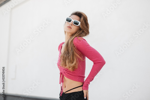 Sexy beautiful woman model hipster with red hair with trendy white sunglasses in a fashionable knitted cropped top with long sleeves and a skirt walking in the city on a white background © alones