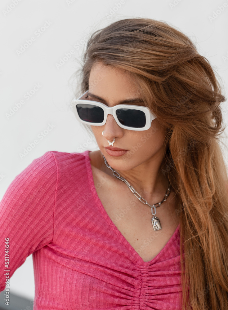 Naklejka premium Cool trendy pretty redhead girl with white fashion sunglasses in a stylish pink knit top walking outside on a sunny summer day