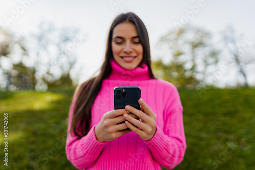 young smiling woman in pink sweater walking in green park using phone