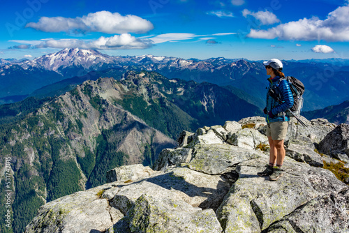 Adventurous athletic female hiker standing on top of a rugged mountain in the Pacific Northwest with jagged mountains in the background. 
