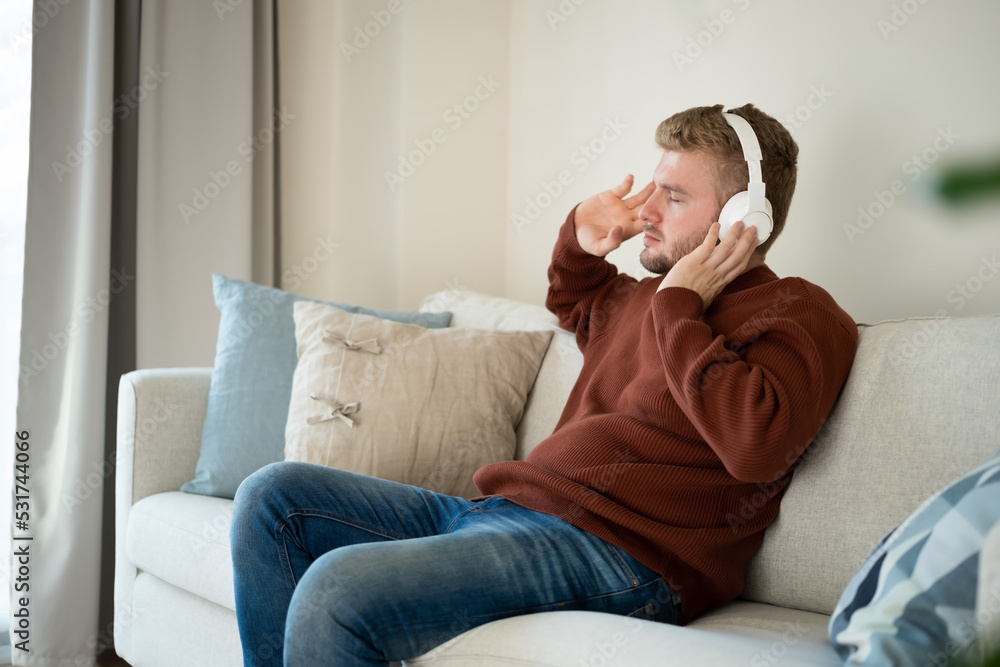 Fototapeta premium Young caucasian blond curly hair man listening to music with big white handsfree headphones sitting on sofa at home on neutral white background. Handsome adult guy portrait lifestyle.Copyspace.