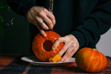 The Halloween Autumn Orange Pumpkin in Men's Hands in a Black Sweater with a kitchen knife prepares for a holiday on a white plate on a table with a checkered plaid