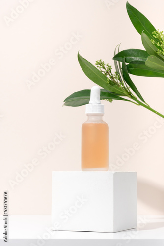 Mockup cosmetics serum with a pipette in a transparent frosted glass and orange liquid. Facial peeling with lactic Aha acid on a white cubic pedestal, beige background with leaves