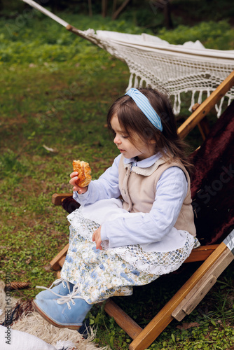 little pretty girl eating pretzel and buns. Little baby of 3 years old in retro vintage dress having fun and smiling in forest outdoors, wraooed with a blanket. Picnic in park photo