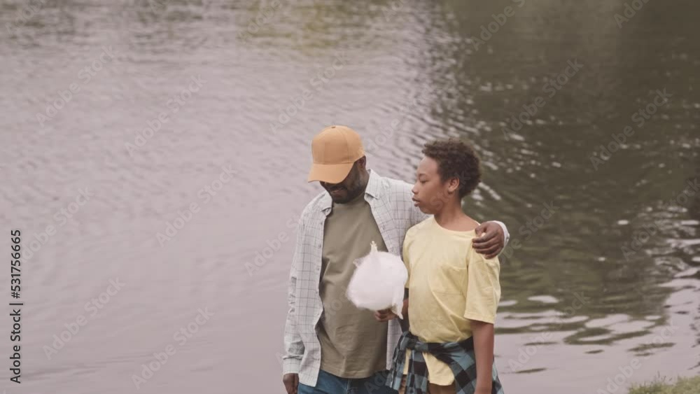 Medium slowmo of happy African American man and his 12 year old son with candy floss strolling along river and chatting, spending day together outdoors Medium slowmo of happy African American man and his 12 year old son with candy floss strolling along river and chatting, spending day together outdoors