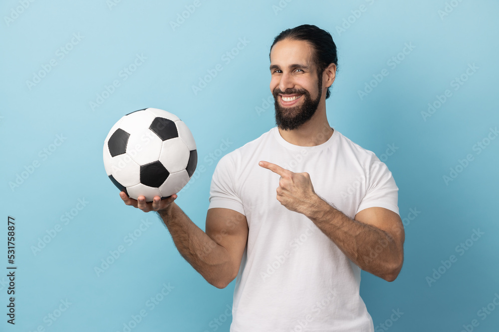 Portrait of delighted man with beard wearing white T-shirt pointing at soccer ball in hand and looking at camera with toothy smile. Indoor studio shot isolated on blue background. Portrait of delighted man with beard wearing white T-shirt pointing at soccer ball in hand and looking at camera with toothy smile. Indoor studio shot isolated on blue background.