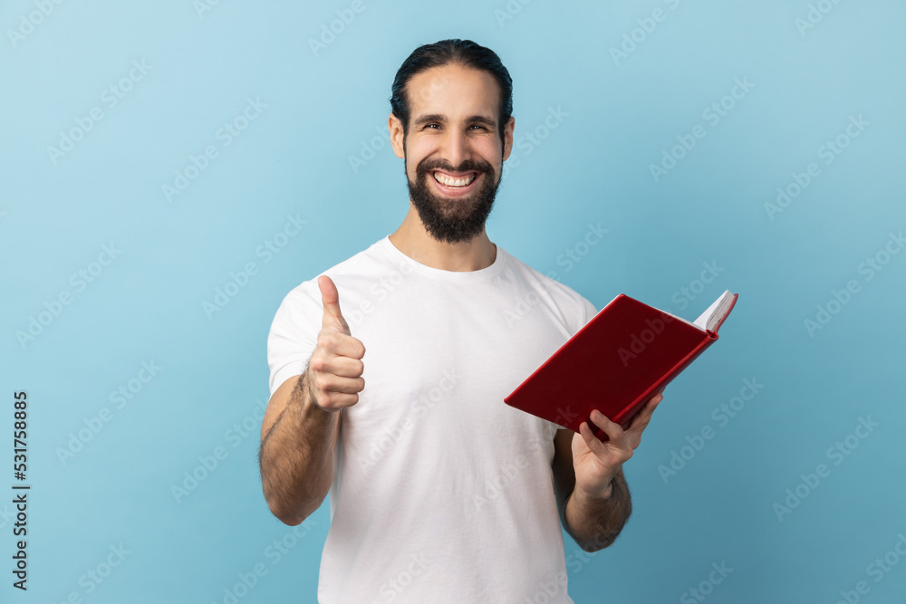 Portrait of positive happy man with beard wearing white T-shirt showing thumbs up gesture holding and reading book, likes genre and plot. Indoor studio shot isolated on blue background. Portrait of positive happy man with beard wearing white T-shirt showing thumbs up gesture holding and reading book, likes genre and plot. Indoor studio shot isolated on blue background.