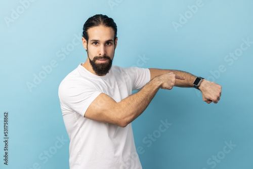 Portrait of impatient angry man with beard wearing white T-shirt pointing at wrist watch and expressing dissatisfaction with late time, delayed meeting. Indoor studio shot isolated on blue background.
