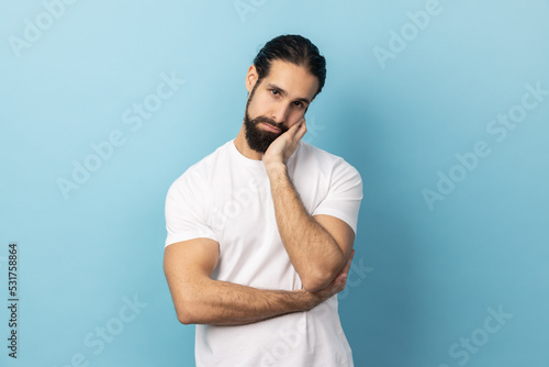 Portrait of man with beard wearing white T-shirt leaning head on hand looking at camera with indifferent look, laziness and apathy, procrastination. Indoor studio shot isolated on blue background.