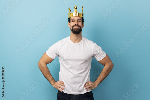 Portrait of confident man with beard wearing white T-shirt and gold crown, standing with hands on hips, looking at camera with conceited expression. Indoor studio shot isolated on blue background.