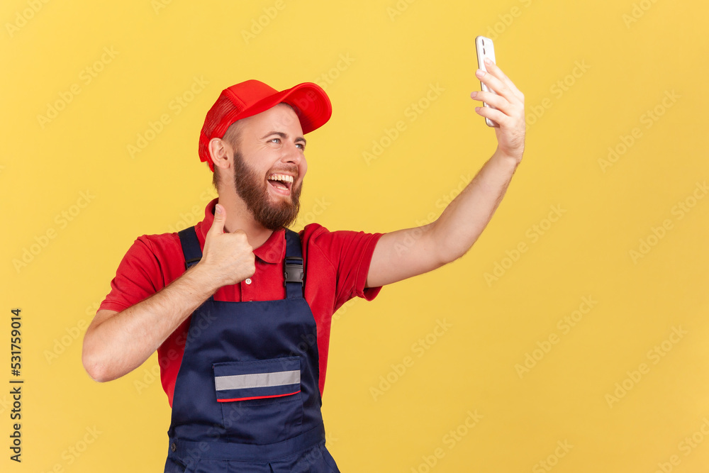 Portrait of positive worker in overalls taking selfie or talking on video call, looking at device camera and showing thumb up to client. Indoor studio shot isolated on yellow background.