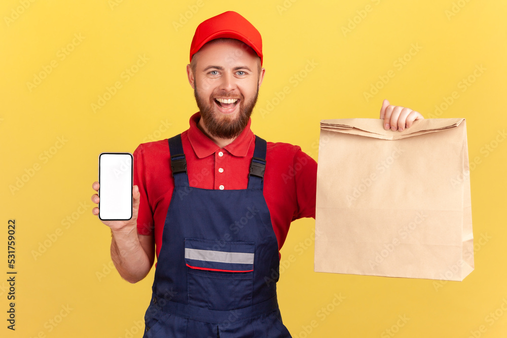 Joyful satisfied deliveryman holding paper parcel and showing smart phone with blank display for advertisement, looking at camera with smile. Indoor studio shot isolated on yellow background.