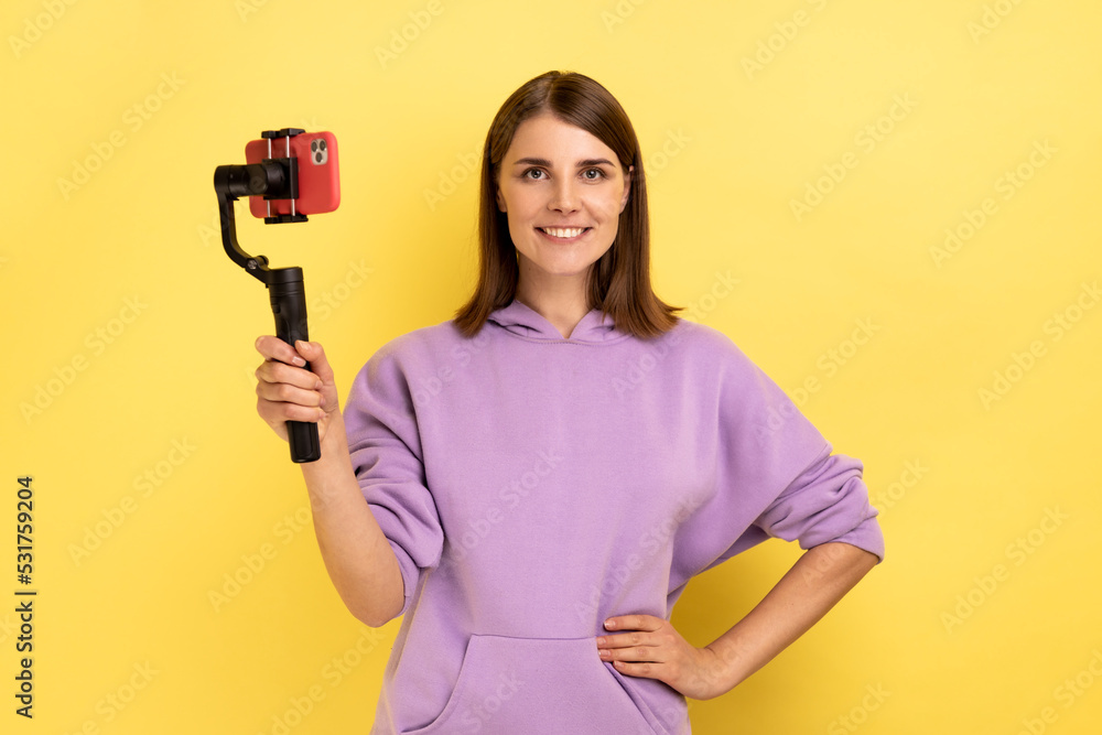 Portrait of delighted woman using cell phone and steadicam for broadcasting livestream, keeping hand on hips, wearing purple hoodie. Indoor studio shot isolated on yellow background. Portrait of delighted woman using cell phone and steadicam for broadcasting livestream, keeping hand on hips, wearing purple hoodie. Indoor studio shot isolated on yellow background.