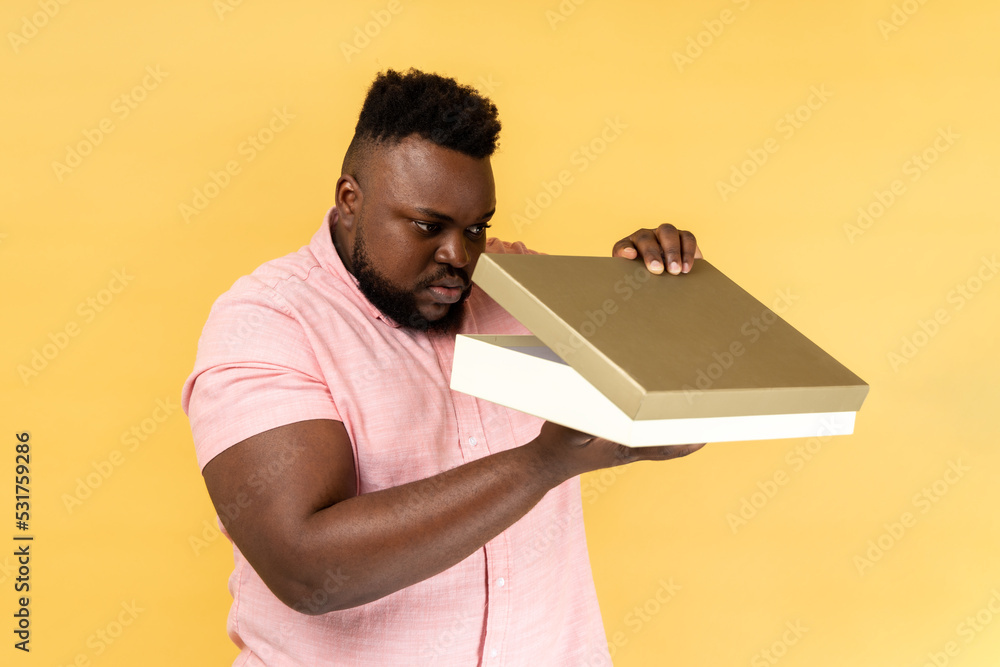 Portrait of curious serious bearded young adult man wearing pink shirt opening gift box and peeping inside, displeased with present. Indoor studio shot isolated on yellow background. Portrait of curious serious bearded young adult man wearing pink shirt opening gift box and peeping inside, displeased with present. Indoor studio shot isolated on yellow background.