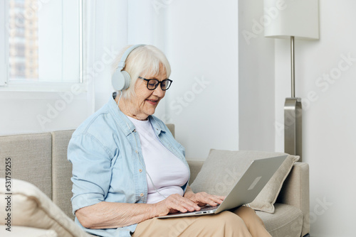 a happy, joyful old lady is sitting on a beige sofa with glasses on her face and headphones on her head working at home in a comfortable environment learning new things