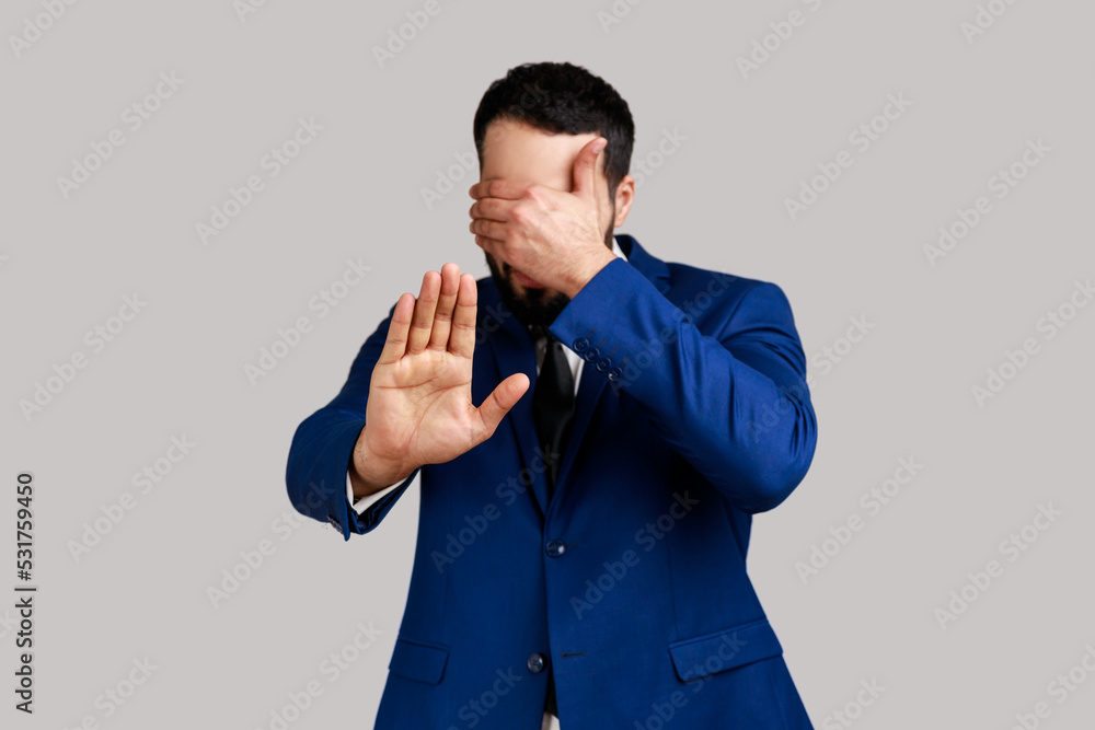 Man closing eyes with hand, outstretching palm, dont want to see that, ignoring problems, hiding from stressful situations, wearing official style suit. Indoor studio shot isolated on gray background. Man closing eyes with hand, outstretching palm, dont want to see that, ignoring problems, hiding from stressful situations, wearing official style suit. Indoor studio shot isolated on gray background.