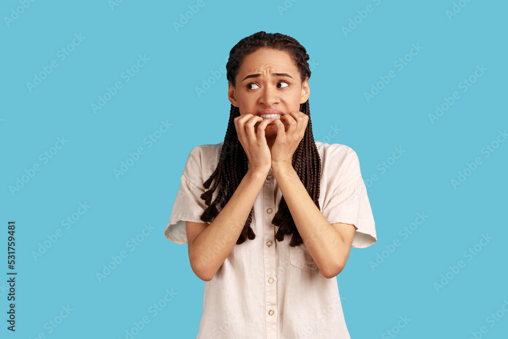 Portrait of puzzled woman with black dreadlocks has worried nervous expression, keeps hand on mouth, hears unexpected bad news, wearing white shirt. Indoor studio shot isolated on blue background. Portrait of puzzled woman with black dreadlocks has worried nervous expression, keeps hand on mouth, hears unexpected bad news, wearing white shirt. Indoor studio shot isolated on blue background.