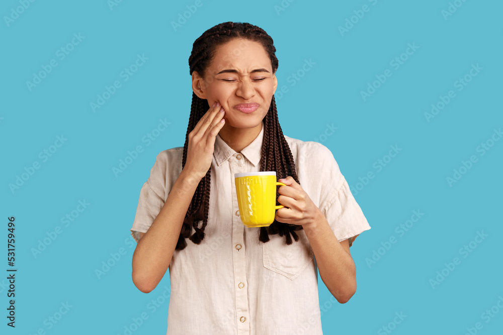 Portrait of unhealthy woman with black dreadlocks wearing white shirt. holding cup in hands, having tooth pain after drinking cold or hot beverage. Indoor studio shot isolated on blue background. Portrait of unhealthy woman with black dreadlocks wearing white shirt. holding cup in hands, having tooth pain after drinking cold or hot beverage. Indoor studio shot isolated on blue background.