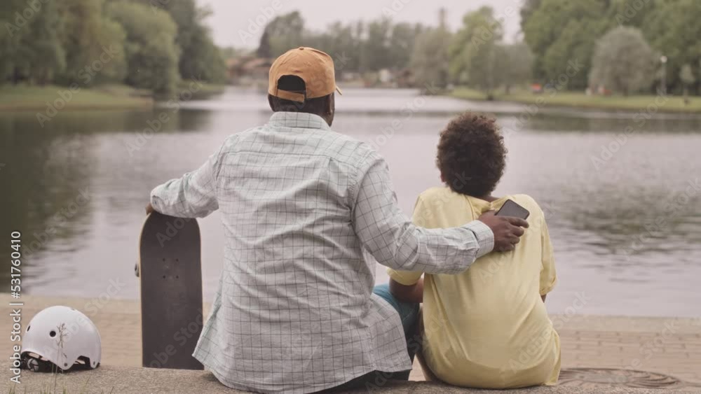 Rear view slowmo of Black man with teenage son sitting by river enjoying nature and each other company Rear view slowmo of Black man with teenage son sitting by river enjoying nature and each other company
