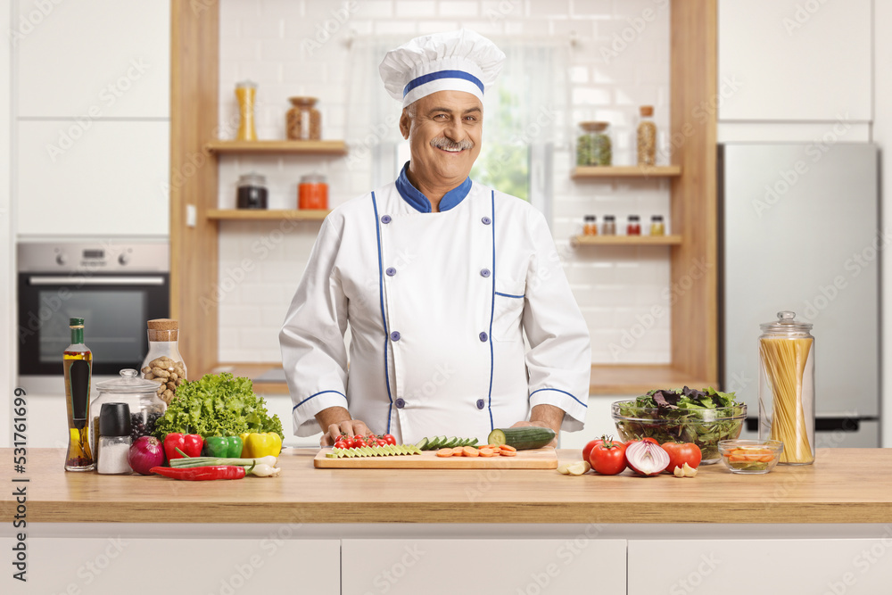 Smiling mature male chef posing behing a counter with vegetables in a kitchen