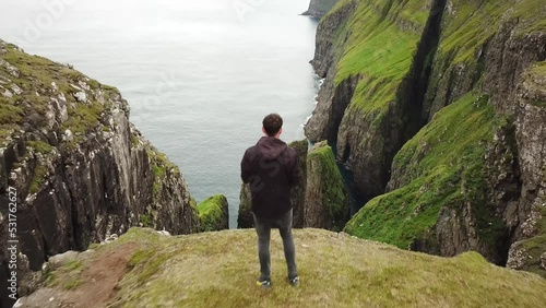 4K Aerial drone view of a man looking down to Dunnesdrangar, Faroe Islands. Amazing nature, huge cliffs and mountains in Faroe Islands. Vagar Island. Many flying sea birds. Sunny day in summer.