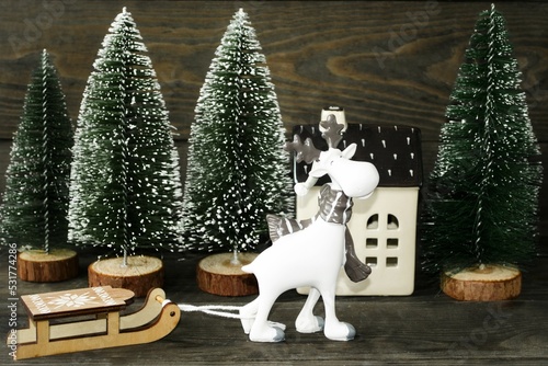 Decorative card for New Year and Christmas holiday.  A beautiful, cute deer with a wooden sleigh against the background of green Christmas trees and a house.  Concept winter holiday atmosphere.