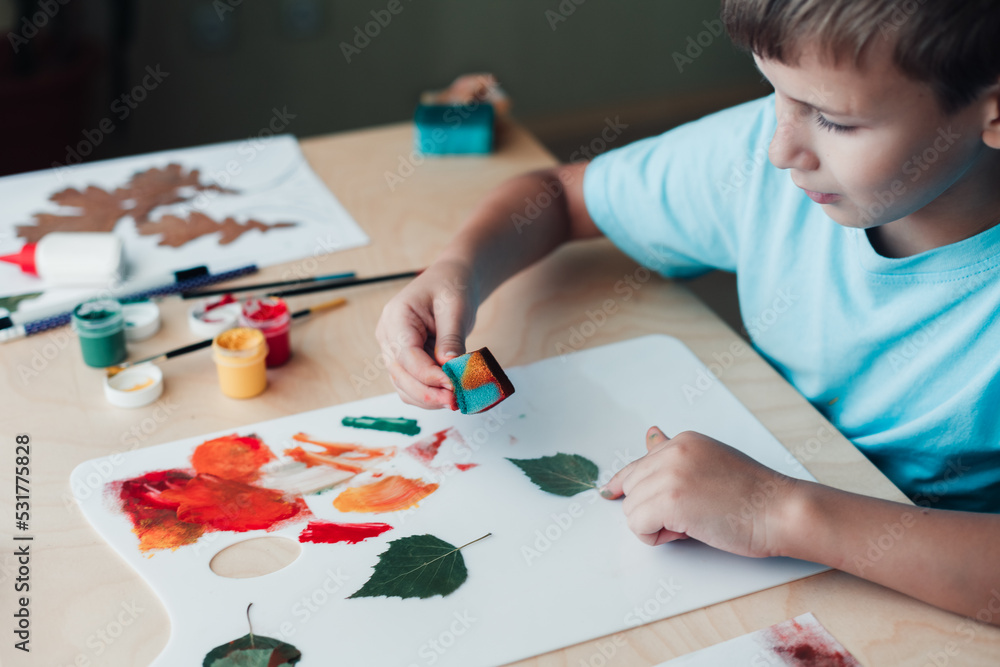 Cute child sitting at desk and making picture from dry birch leaves Cute child sitting at desk and making picture from dry birch leaves