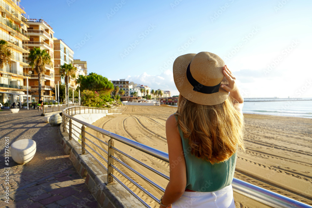 Tourism in Calabria. Back view of girl holding straw hat walking on Crotone promenade on Calabria coast, Italy.