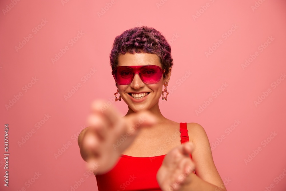 Fashion portrait of a woman with a short haircut in colored sunglasses with unusual accessories with earrings smiles on a pink bright background shows her hands to the camera Fashion portrait of a woman with a short haircut in colored sunglasses with unusual accessories with earrings smiles on a pink bright background shows her hands to the camera