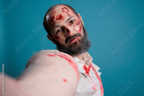 Eerie zombie holding camera to take picture, looking creepy and dangerous over blue background. Scary brain eating devil with horror look and aggressive bloody face taking frightening photo.