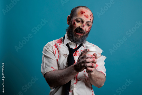 Dangerous zombie looking at cup of coffee, wanting to dirnk beverage and being aggressive in studio. Scary evil monster holding cup and having bloody wounds, apocalyptic eerie devil. photo