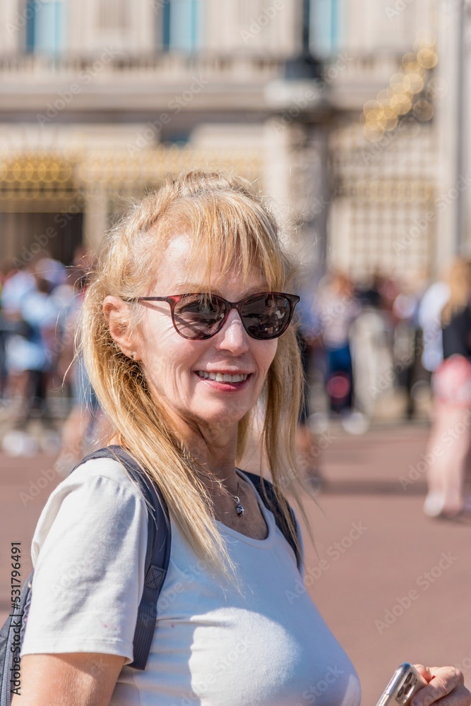 Portrait of a mature blonde woman with sunglasses looking at the camera. Unfocused Buckingham Palace and people behind.
