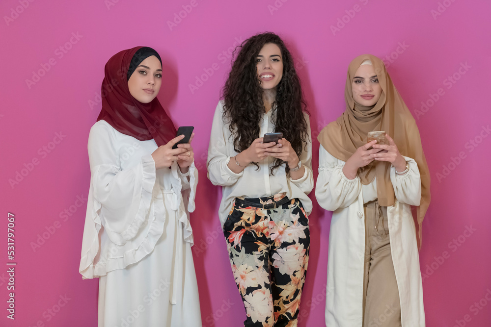 Group of beautiful muslim women two of them in fashionable dress with hijab using mobile phone isolated on pink background representing modern islam fashion technology and ramadan kareem concept