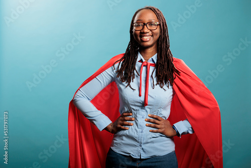 Proud superhero woman with superpower abilities wearing mighty hero cloak while smiling confident at camera. Joyful happy smiling young adult person posing as justice defender on blue background. photo
