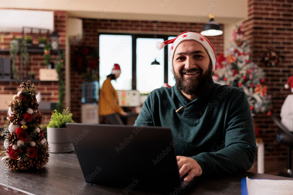 Portrait of office employee working on company laptop and sitting in office decorated with christmas tree and lights, festive ornaments. Smiling man wearing santa hat during seasonal xmas time. Portrait of office employee working on company laptop and sitting in office decorated with christmas tree and lights, festive ornaments. Smiling man wearing santa hat during seasonal xmas time.