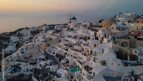 Cinematic Aerial Shot of Oia Village, Santorini, Greece - Cyclades - close-up on famous Windmills silhouetting against the Aegean Sea, while tourists enjoy sunset from typical whitewashed houses