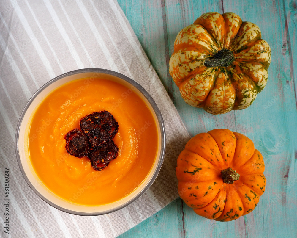 pumpkin soup puree in a round plate with slices of dried tomato and two small pumpkins stand side by side on a blue wooden table. view from above. vegan pumpkin soup puree in a round plate with slices of dried tomato and two small pumpkins stand side by side on a blue wooden table. view from above. vegan