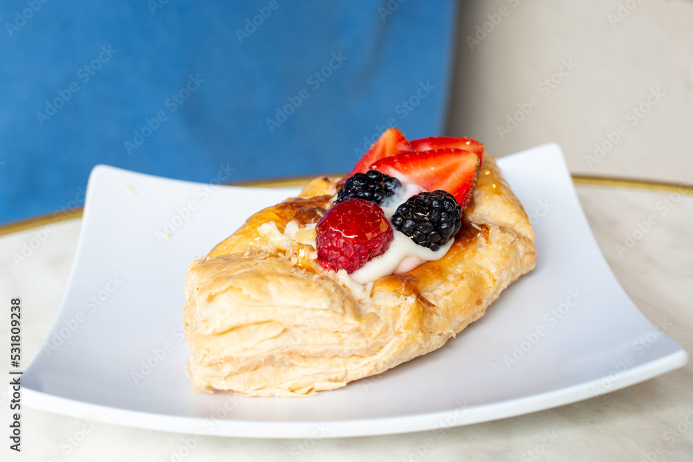 A square fruit filled French puff pastry on a white square plate. The cream and custard filled dessert have strawberries, kiwi, and blackberries on top with a sugar glaze. The pastry is flaky.	