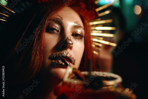 Woman with skeleton makeup at a Halloween party drinks cocktail and looks at camera. All Saints' Night. Celebration in a nightclub. Female in creepy costume of Mexican goddess of death.
